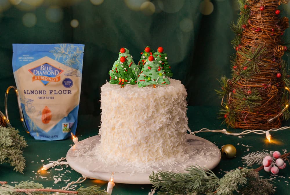 A white frosted cake with coconut flakes and green Christmas tree decorations sits on a plate next to a bag of almond flour and festive holiday decor.