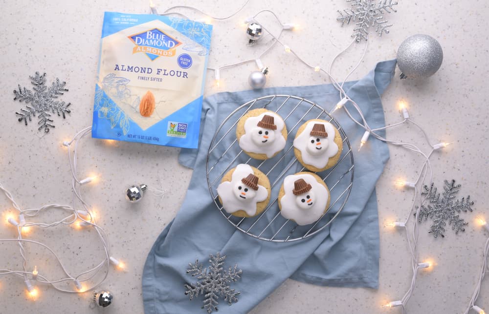 A package of almond flour and four snowman cookies are arranged on a light surface with string lights, silver snowflakes, and a silver ornament.