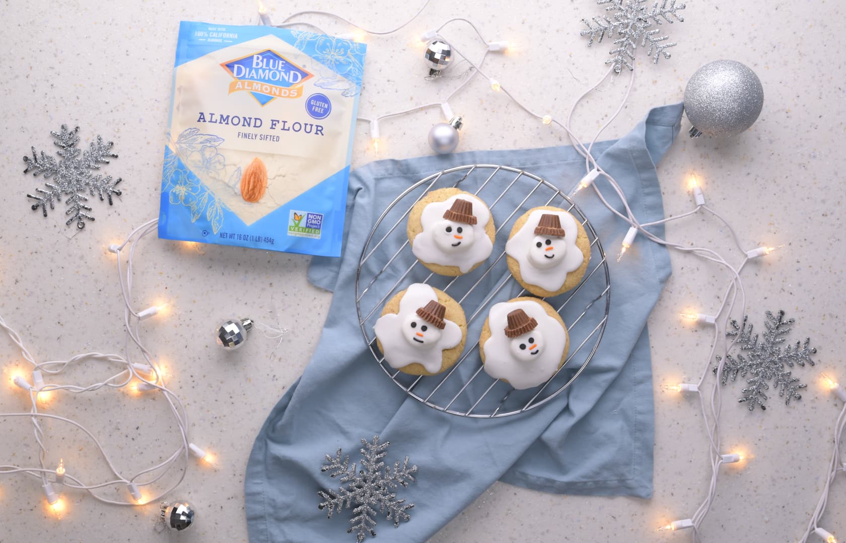 A package of almond flour and four snowman cookies are arranged on a light surface with string lights, silver snowflakes, and a silver ornament.