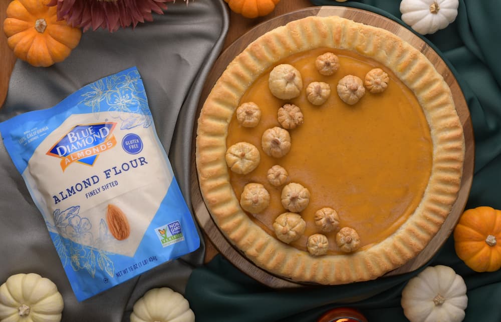 A pumpkin pie with small pastry decorations is on a wooden board surrounded by mini pumpkins and a bag of Blue Diamond almond flour.