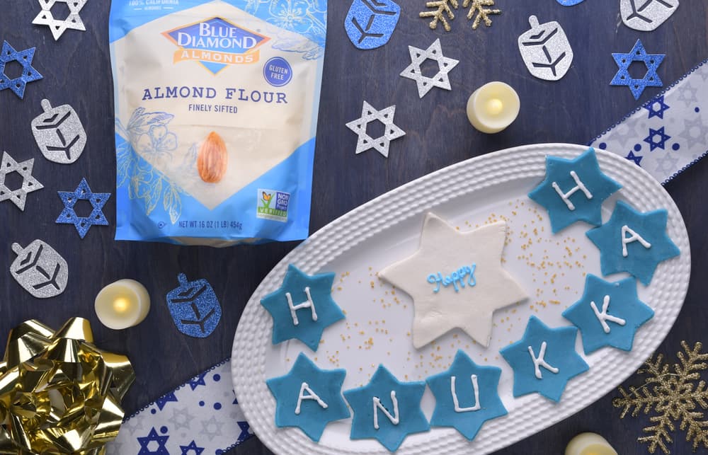A plate of blue star-shaped cookies spelling HANUKKA with a white star cookie, almond flour bag, Hanukkah decorations, and candles on a dark surface.