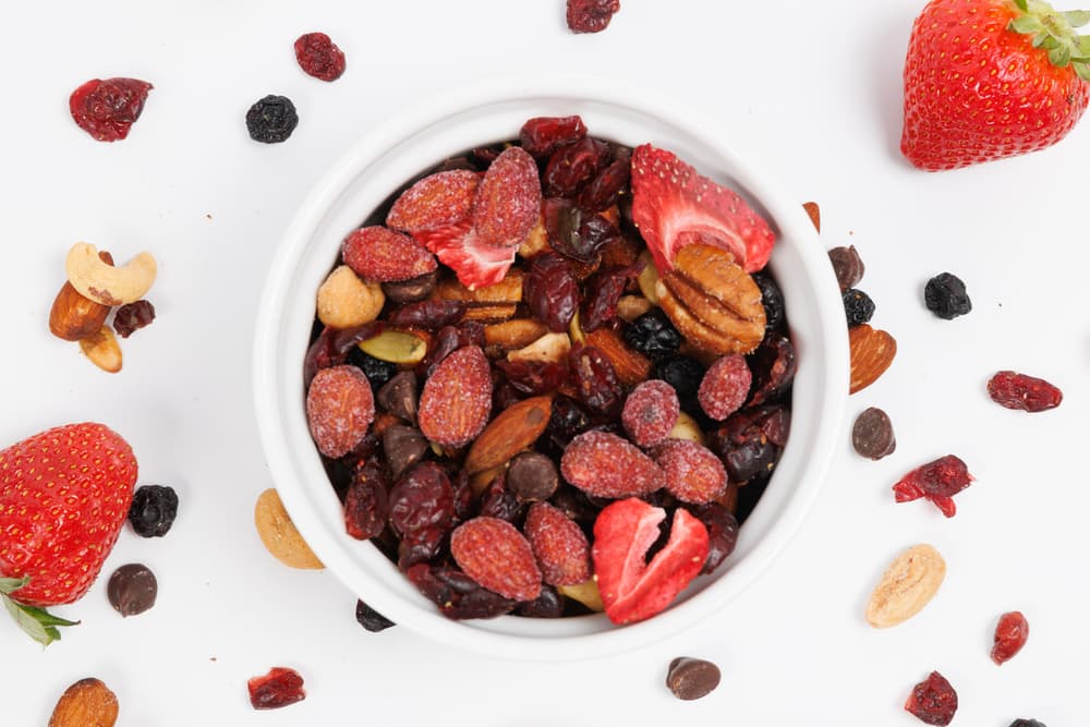 A white bowl filled with mixed nuts, dried berries, and chocolate chips, surrounded by scattered nuts, berries, and fresh strawberries on a white background.