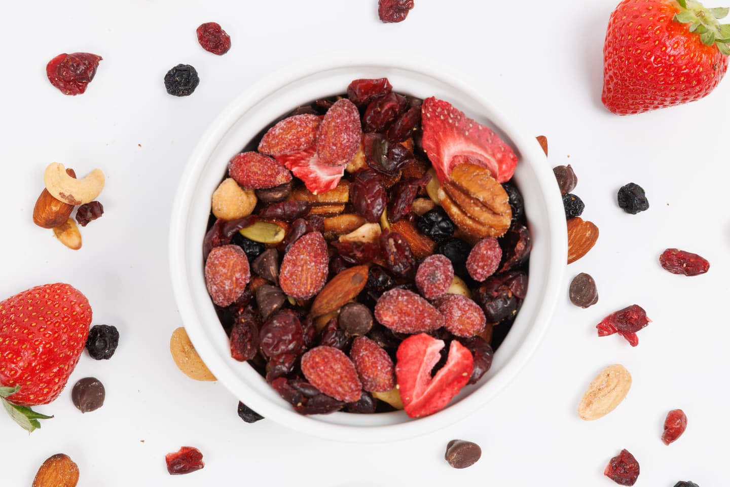 A white bowl filled with mixed nuts, dried berries, and chocolate chips, surrounded by scattered nuts, berries, and fresh strawberries on a white background.