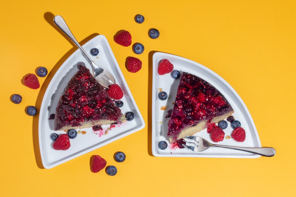 Two slices of berry-topped cheesecake on triangular plates with blueberries and raspberries scattered on a yellow background, each slice with a fork.