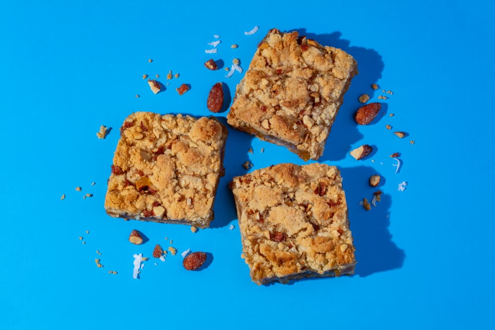 Three square crumb bars with visible nuts and crumbs are arranged on a bright blue surface, with scattered nuts and crumbs surrounding them.