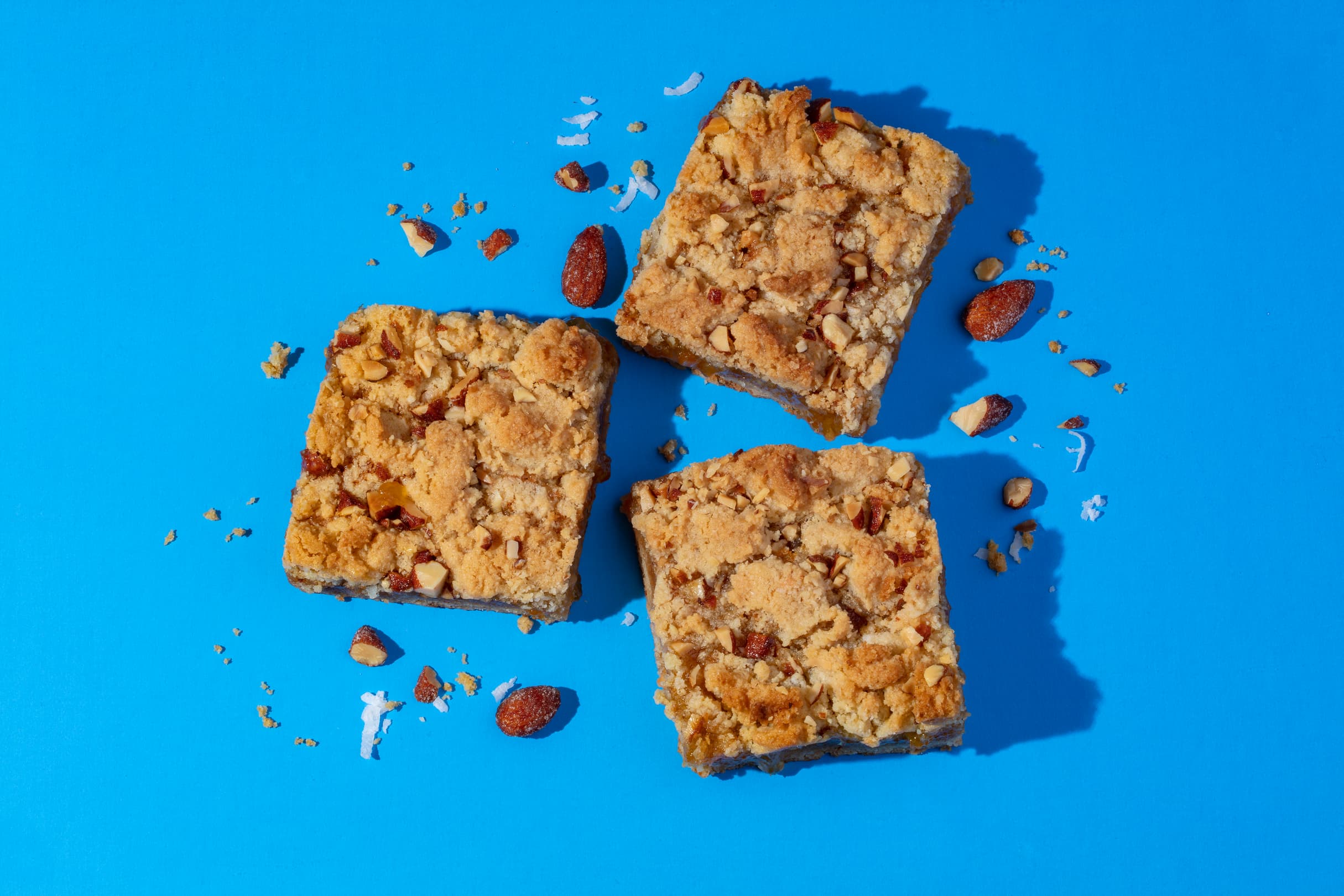 Three square crumb bars with visible nuts and crumbs are arranged on a bright blue surface, with scattered nuts and crumbs surrounding them.