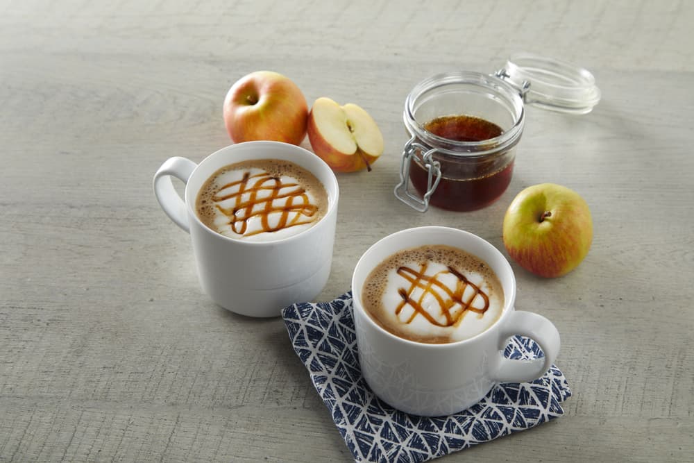 Two mugs of caramel-topped coffee sit on a patterned napkin next to apples, a jar of sauce, and a half apple on a light wooden surface.