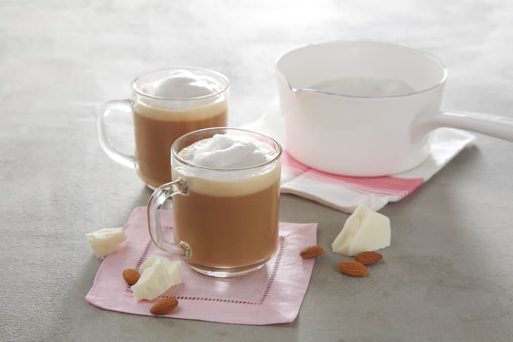 Two glass mugs of frothy coffee sit on pink napkins with almonds and chunks of cocoa butter nearby; a white saucepan is in the background.