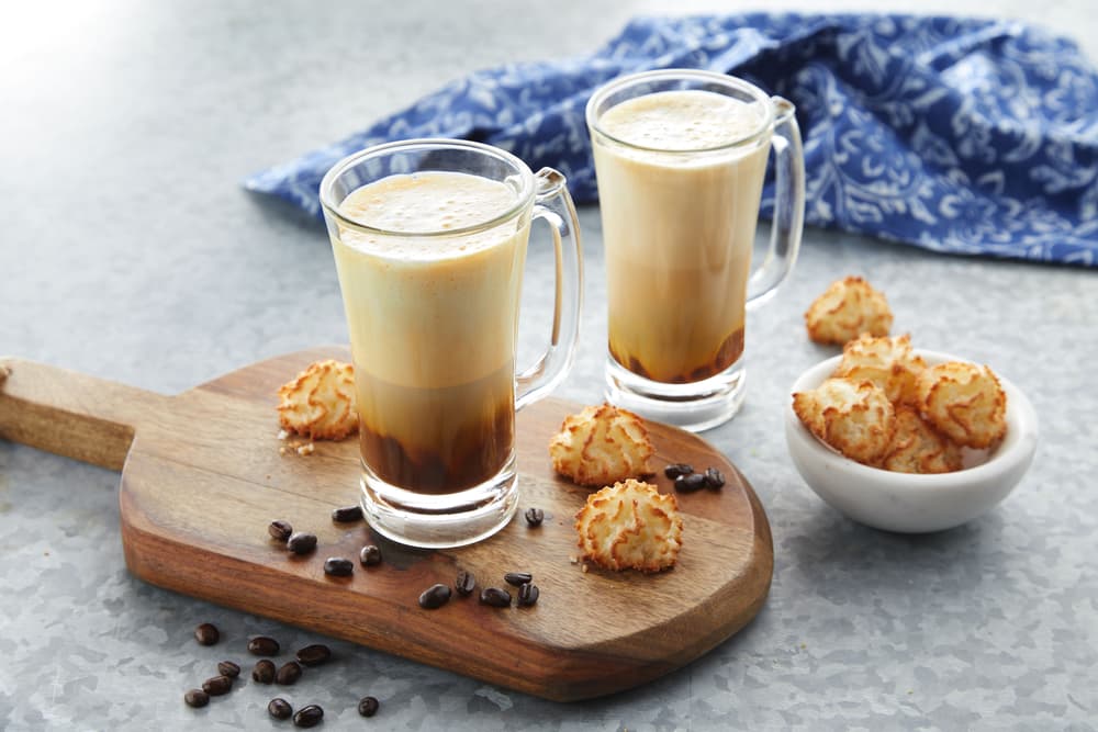 Two glass mugs of layered coffee on a wooden board with coconut macaroons, scattered coffee beans, and a small white bowl of more macaroons on a gray surface. Blue cloth in the background.