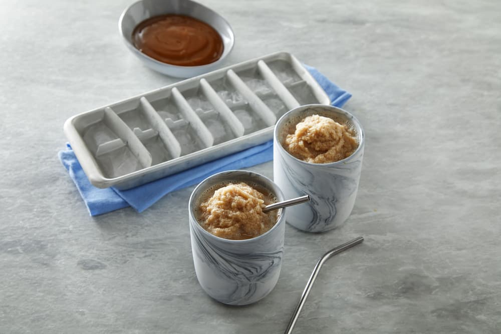Two marble-patterned cups with brown slush and metal straws sit next to an ice cube tray and a dish of caramel sauce on a gray surface.