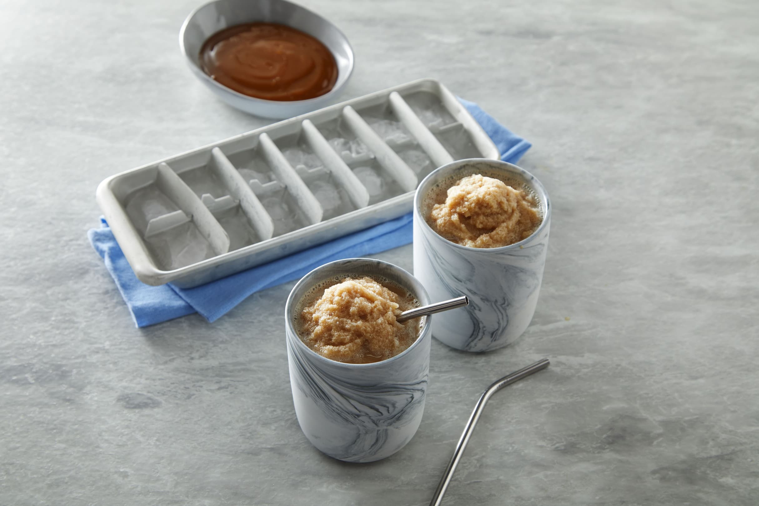 Two marble-patterned cups with brown slush and metal straws sit next to an ice cube tray and a dish of caramel sauce on a gray surface.