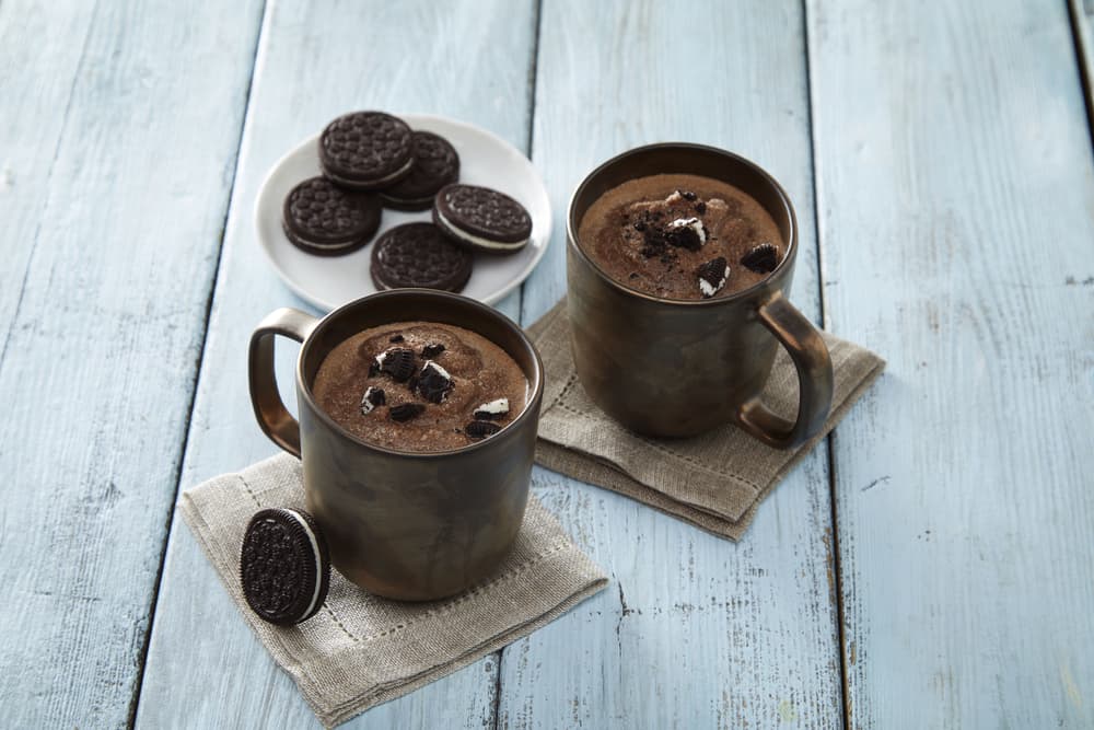 Two mugs of chocolate mug cake with crushed chocolate sandwich cookies on top, placed on napkins beside a plate of whole cookies on a light blue wooden surface.