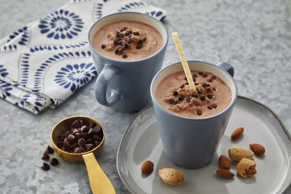 Two blue mugs of chocolate drink topped with chocolate chips, with a golden spoon, almonds, and chocolate chips on a gray plate beside a patterned cloth napkin.