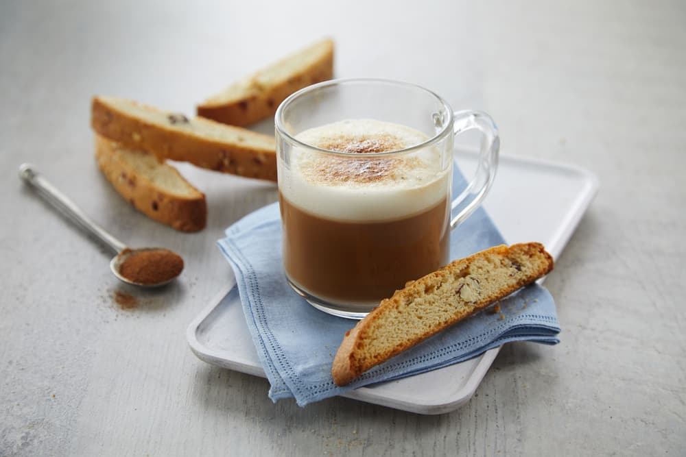 A glass mug of cappuccino sits on a blue napkin with four biscotti and a spoon of cinnamon on a white tray.