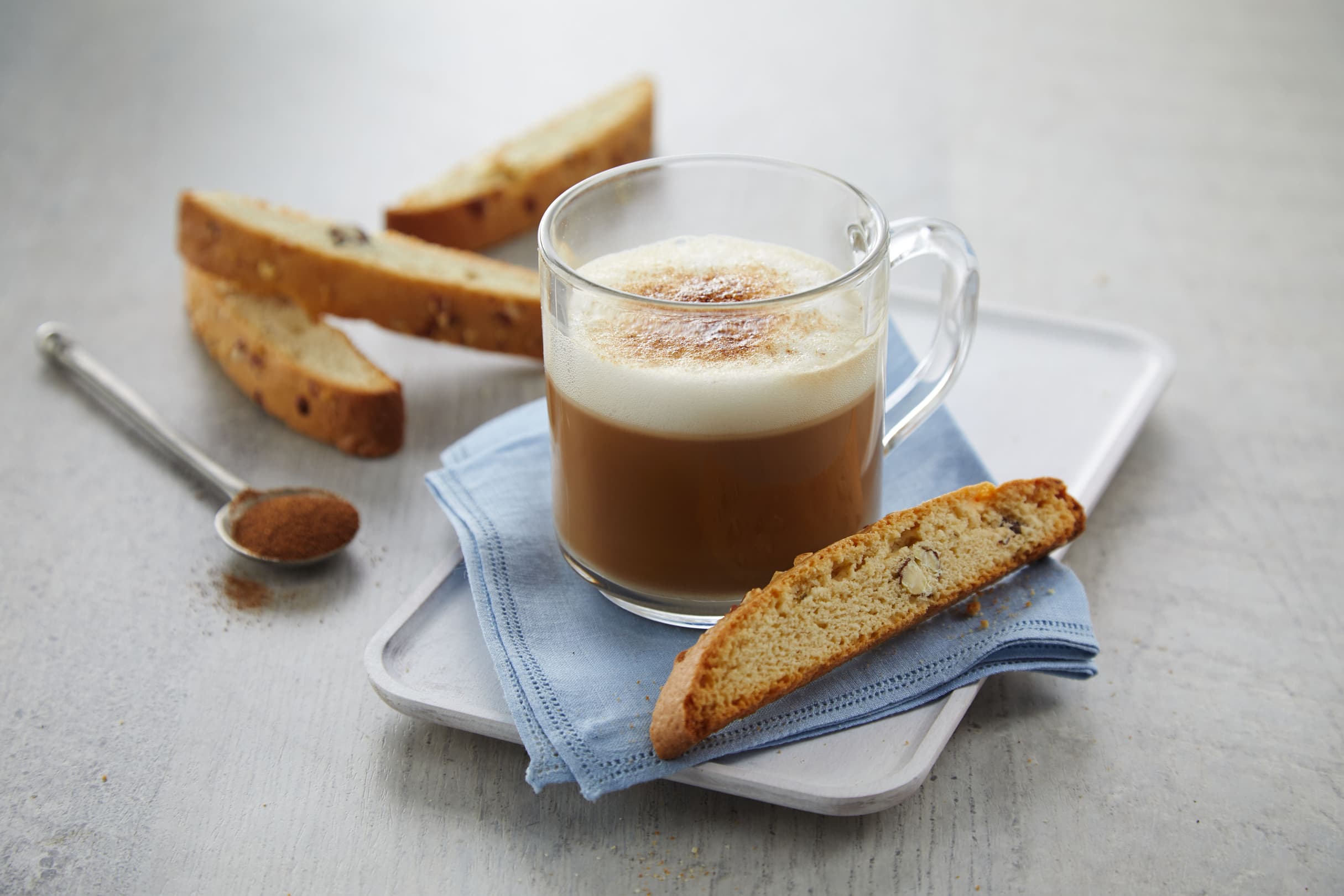 A glass mug of cappuccino sits on a blue napkin with four biscotti and a spoon of cinnamon on a white tray.