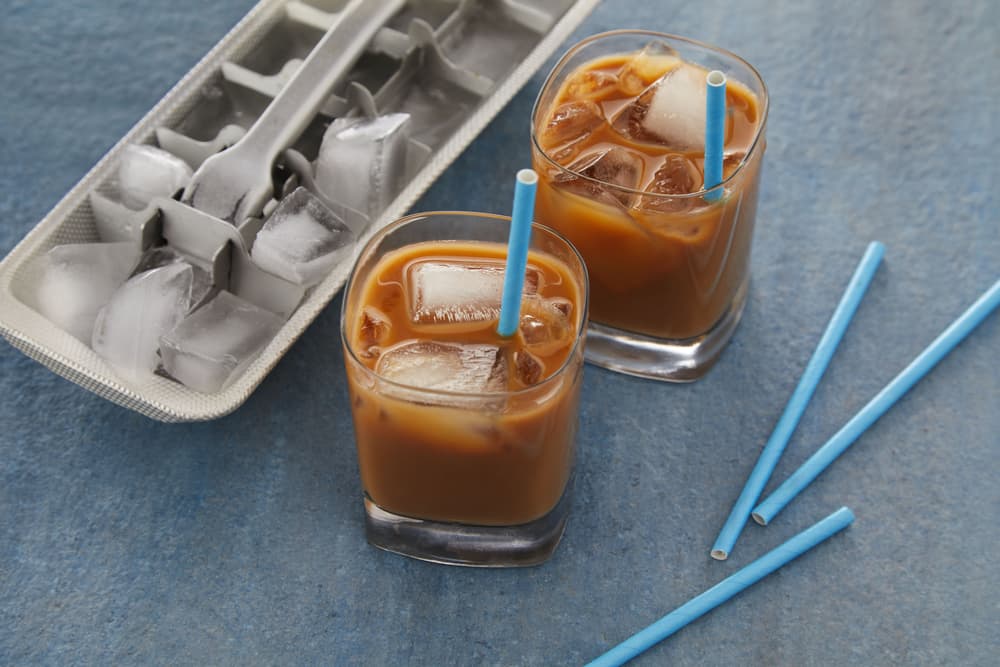Two glasses of iced coffee with blue straws sit next to an ice cube tray and loose blue straws on a blue surface.