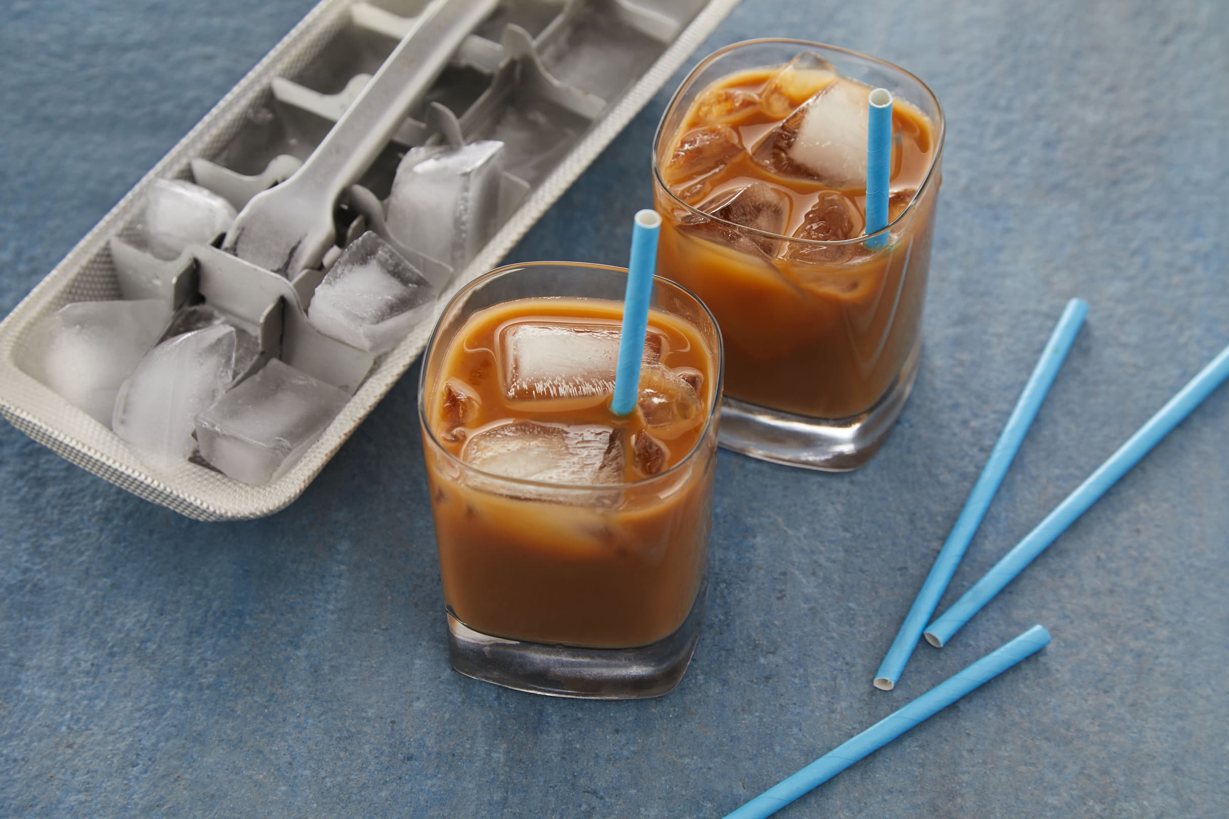 Two glasses of iced coffee with blue straws sit next to an ice cube tray and loose blue straws on a blue surface.