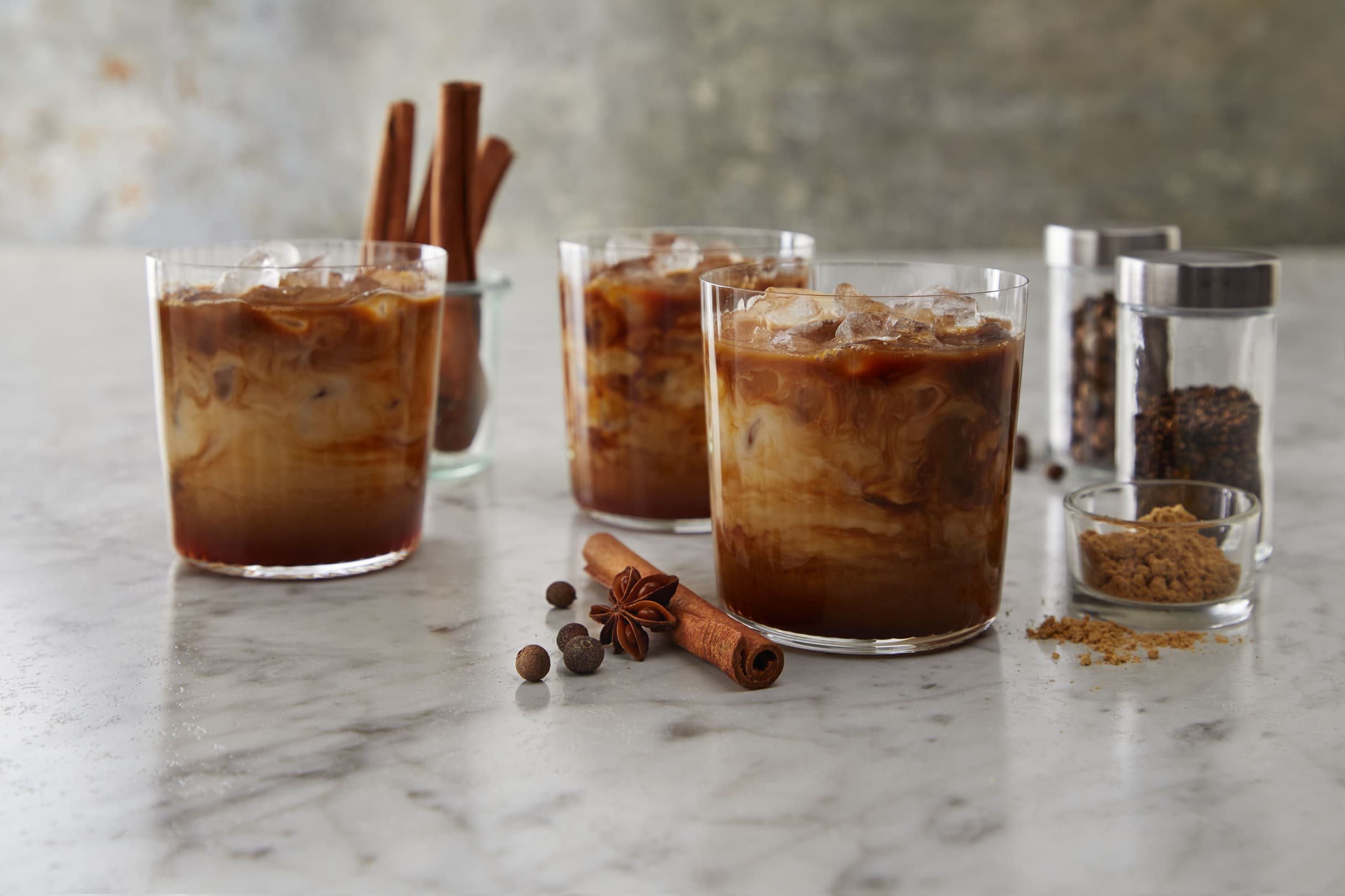 Three iced coffee drinks with milk on a marble surface, surrounded by cinnamon sticks, star anise, whole spices, brown sugar, and spice jars.