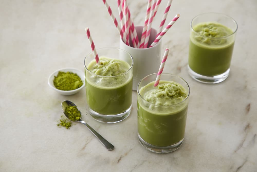 Three glasses of iced matcha latte with pink-striped straws on a marble surface, next to a container of straws and a small dish and spoon with matcha powder.