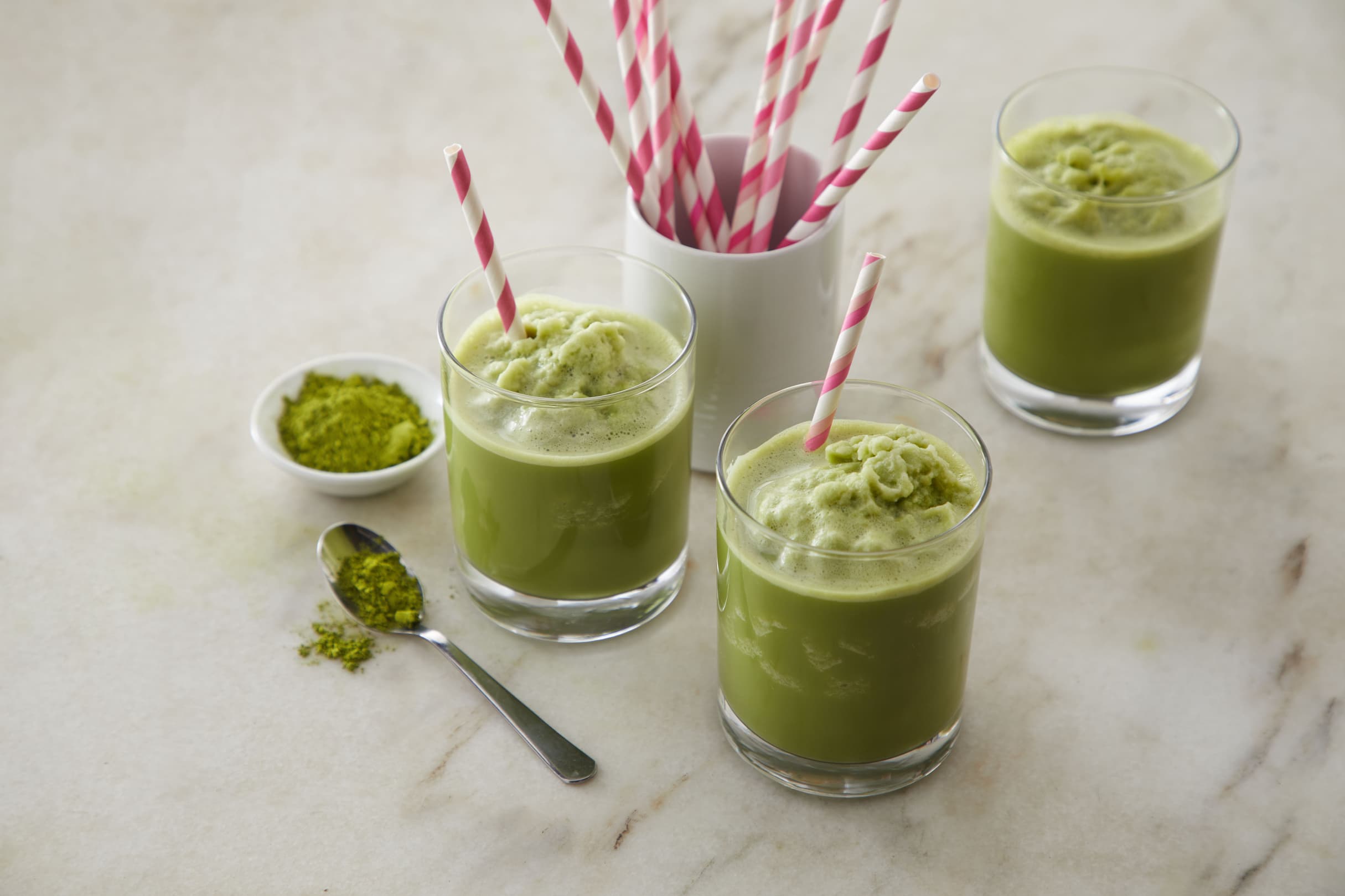 Three glasses of iced matcha latte with pink-striped straws on a marble surface, next to a container of straws and a small dish and spoon with matcha powder.