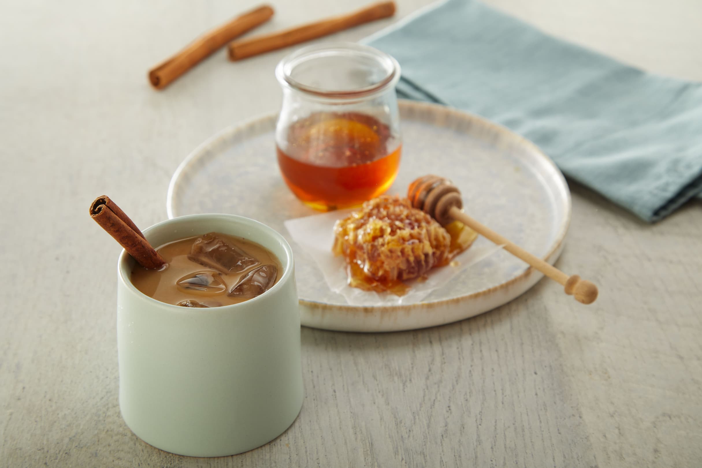 A cup of iced coffee with a cinnamon stick, a plate with honeycomb, a jar of honey, and a honey dipper, next to a blue napkin on a light wooden table.