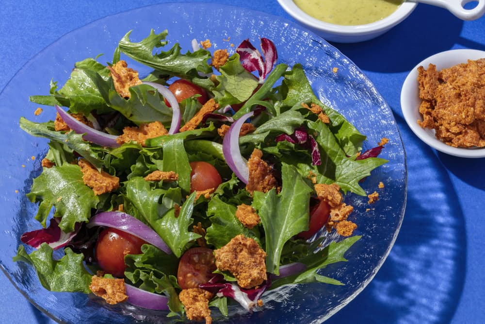A glass bowl of mixed green salad with cherry tomatoes, red onion slices, and crunchy toppings, next to a bowl of dressing and a small dish of croutons on a blue surface.