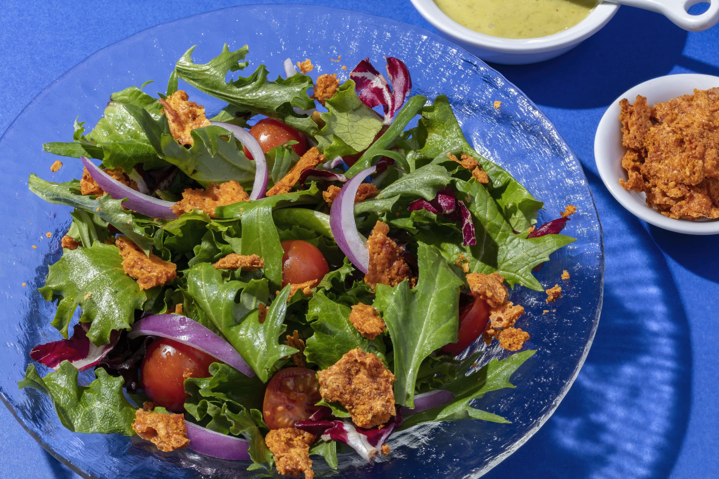 A glass bowl of mixed green salad with cherry tomatoes, red onion slices, and crunchy toppings, next to a bowl of dressing and a small dish of croutons on a blue surface.
