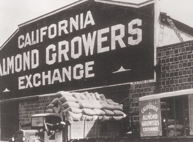 Black-and-white photo of the California Almond Growers Exchange building with sacks of almonds and a truck parked outside.