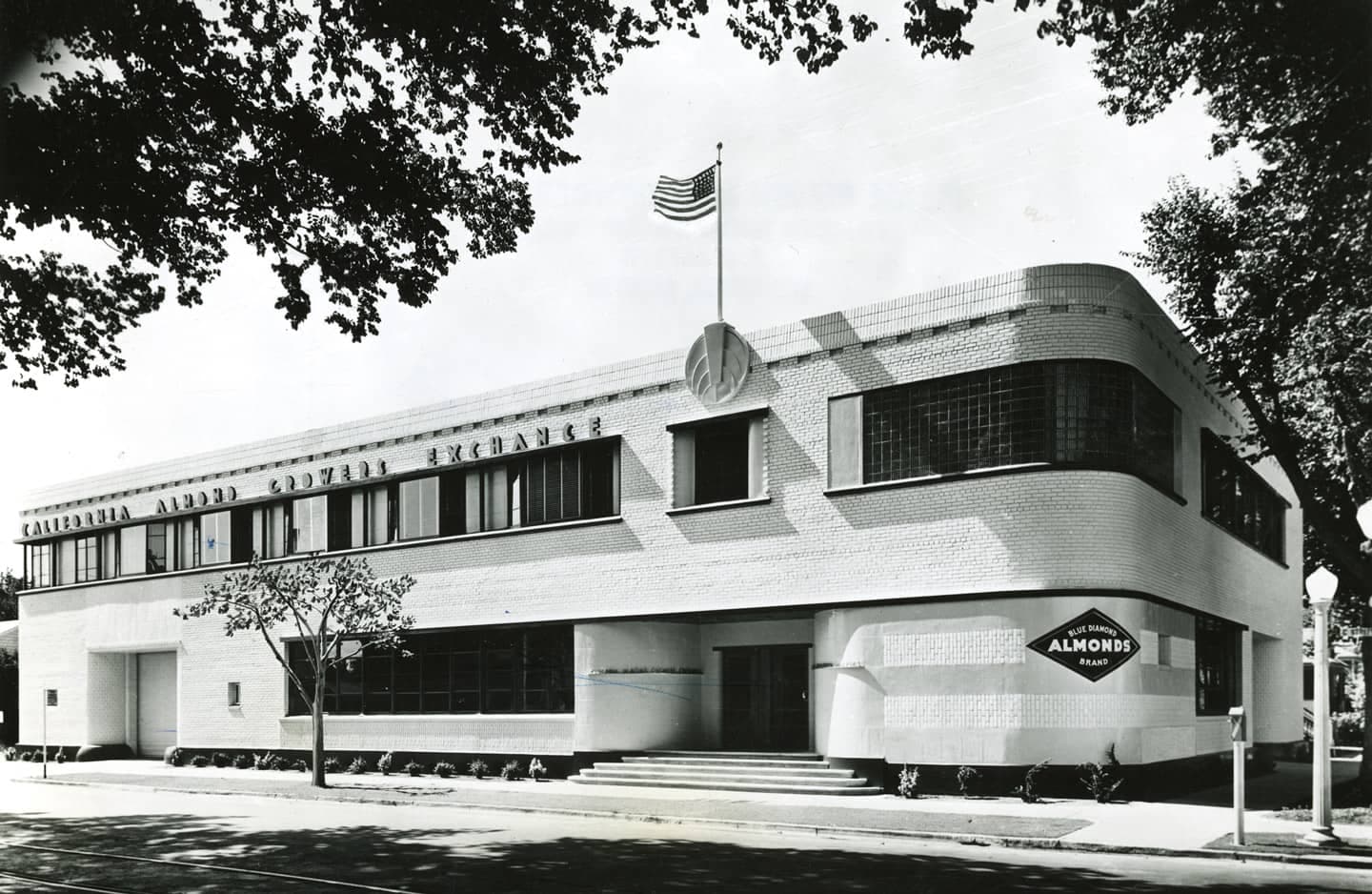 Black-and-white photo of the California Almond Growers Exchange building, a two-story, white Art Deco structure with an American flag and signage, partially shaded by trees.