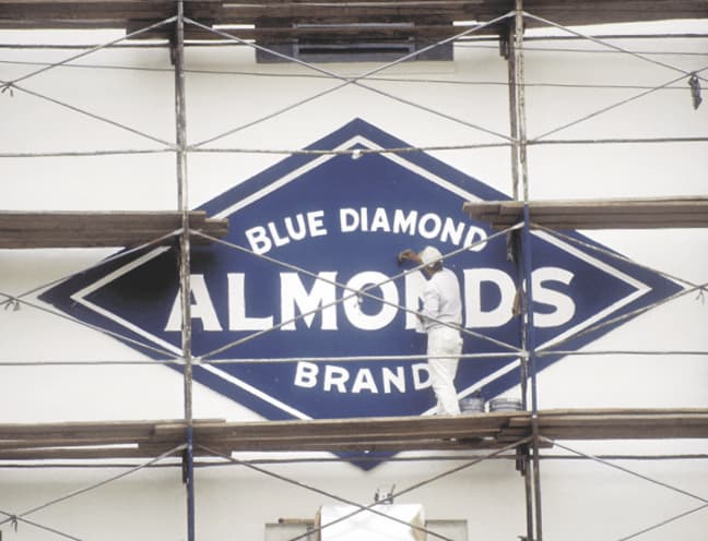 Worker on scaffolding paints a large Blue Diamond Almonds Brand logo on a white wall.
