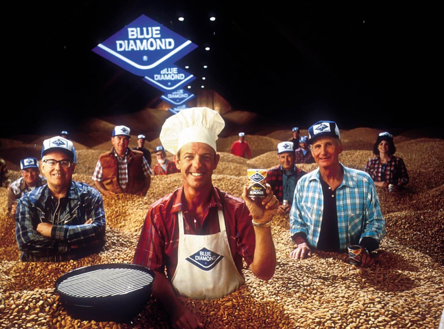 A group of people in Blue Diamond hats and uniforms stand in piles of almonds, with one person in a chefs hat holding a can of Blue Diamond almonds in the foreground.