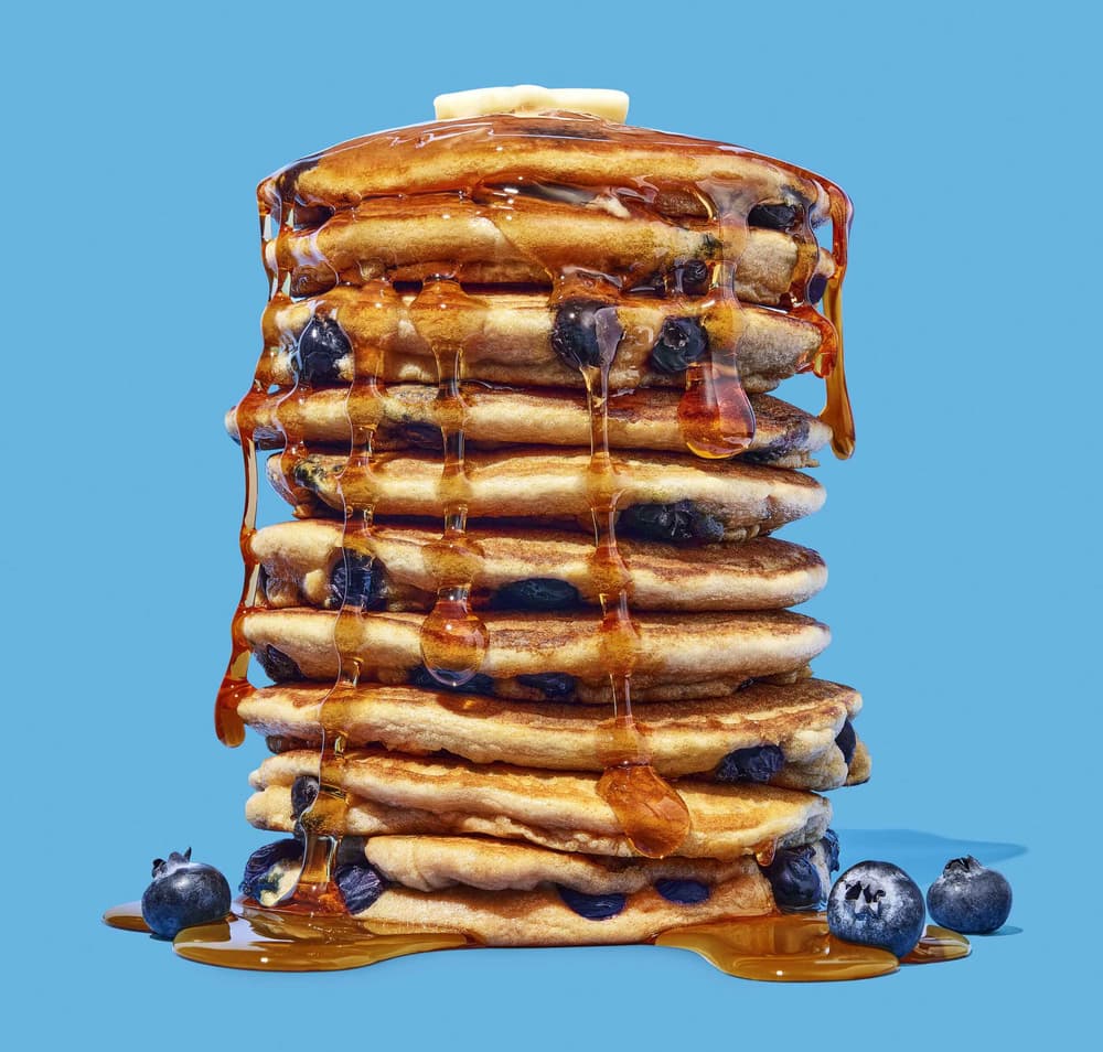A tall stack of blueberry pancakes with syrup drizzling down and a pat of butter on top, set against a solid blue background.