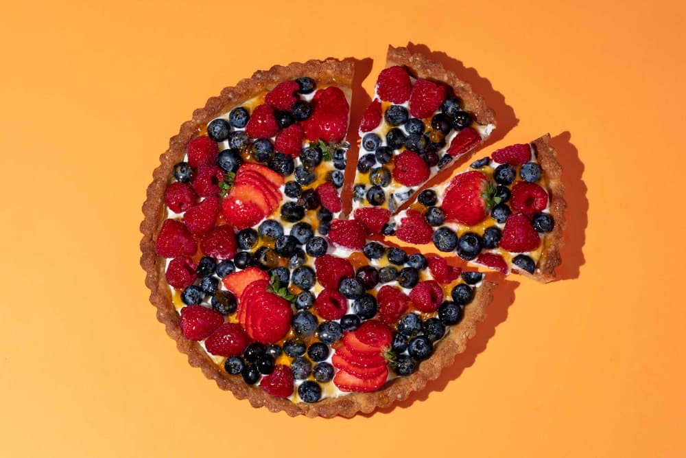 A round fruit tart topped with strawberries, blueberries, raspberries, and kiwi sits on an orange background with two slices slightly separated from the tart.