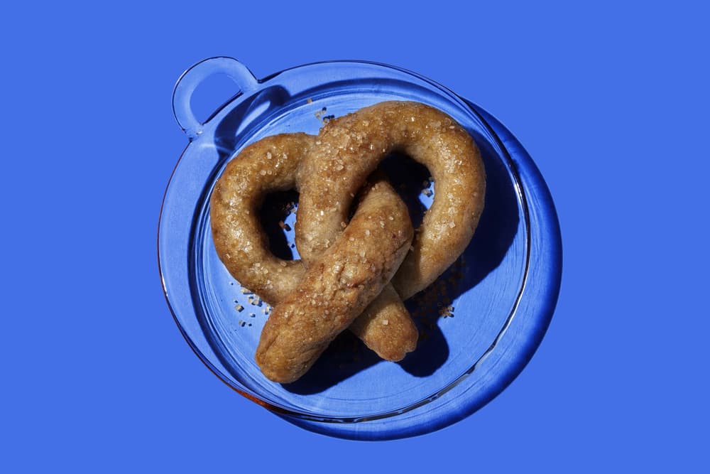A single salted pretzel on a clear glass plate against a solid blue background.