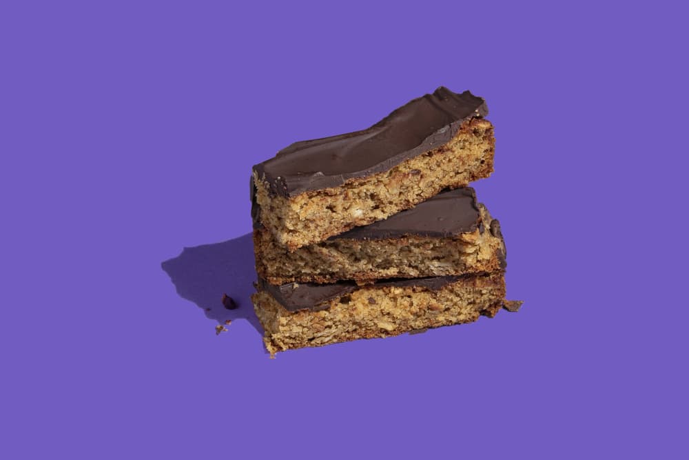 Three chocolate-topped oat bars stacked on top of each other against a solid purple background.