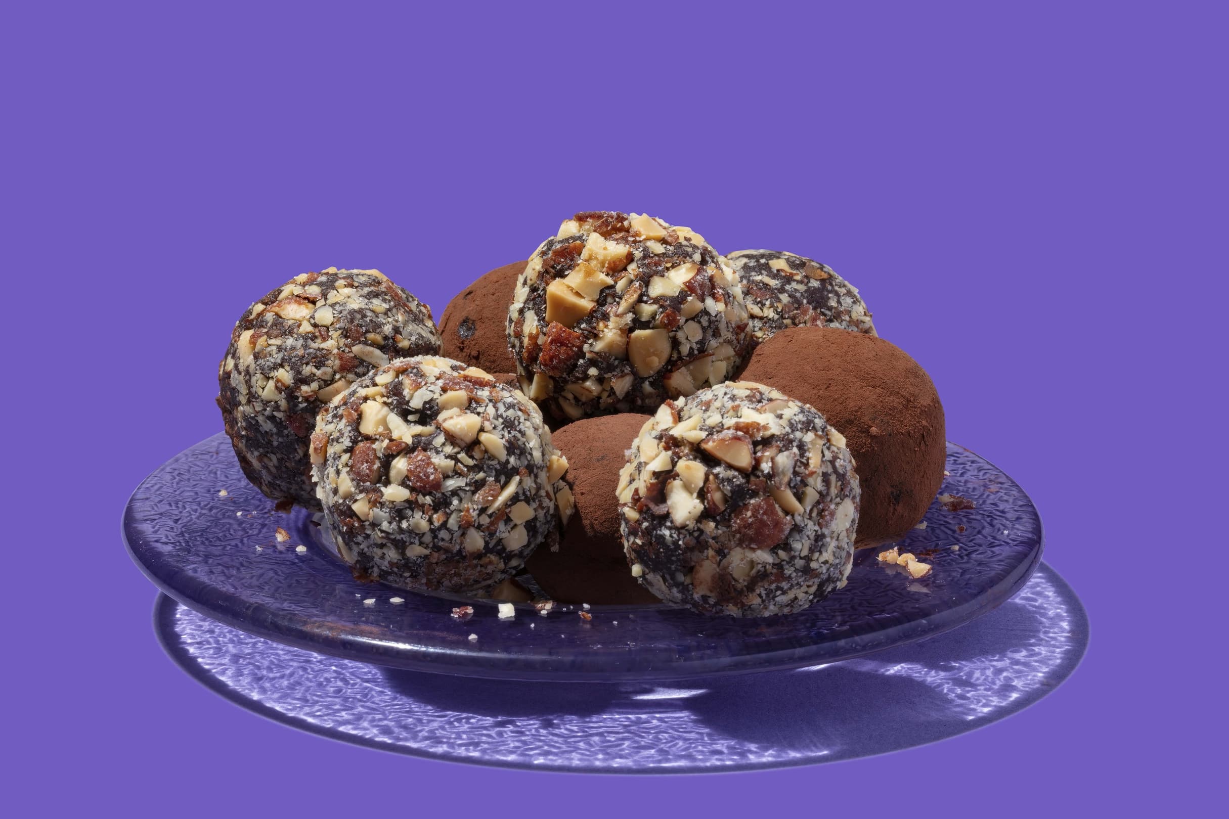 A glass plate with chocolate truffles, some coated in chopped nuts and others in cocoa powder, set against a solid purple background.