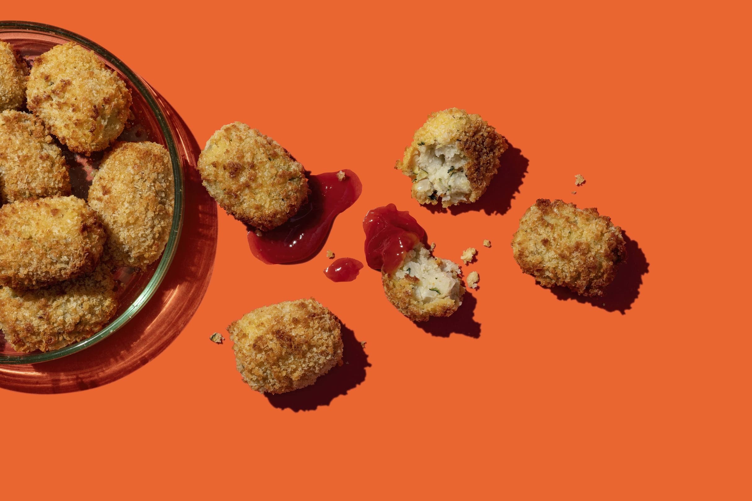 A glass bowl of breaded nuggets sits on an orange surface, with several nuggets and a smear of red dipping sauce beside it.