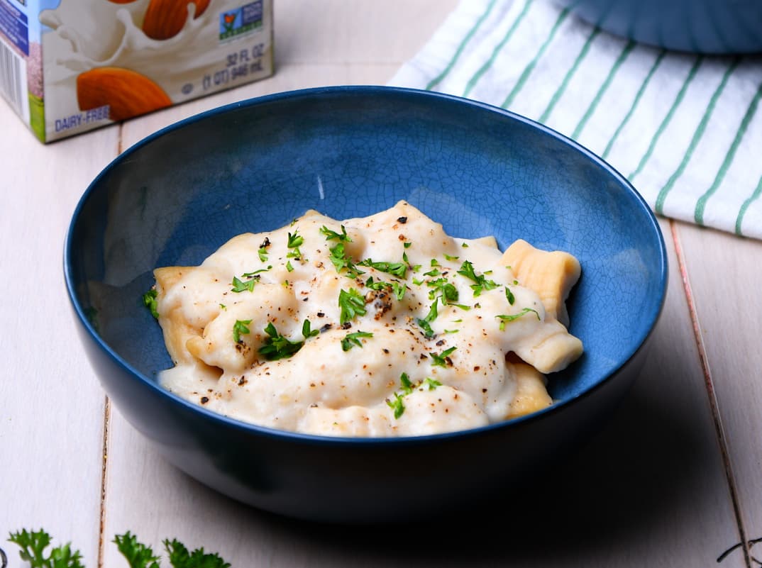 A blue bowl of pasta topped with a creamy white sauce, chopped herbs, and black pepper sits on a light wooden surface next to a striped cloth and a carton.