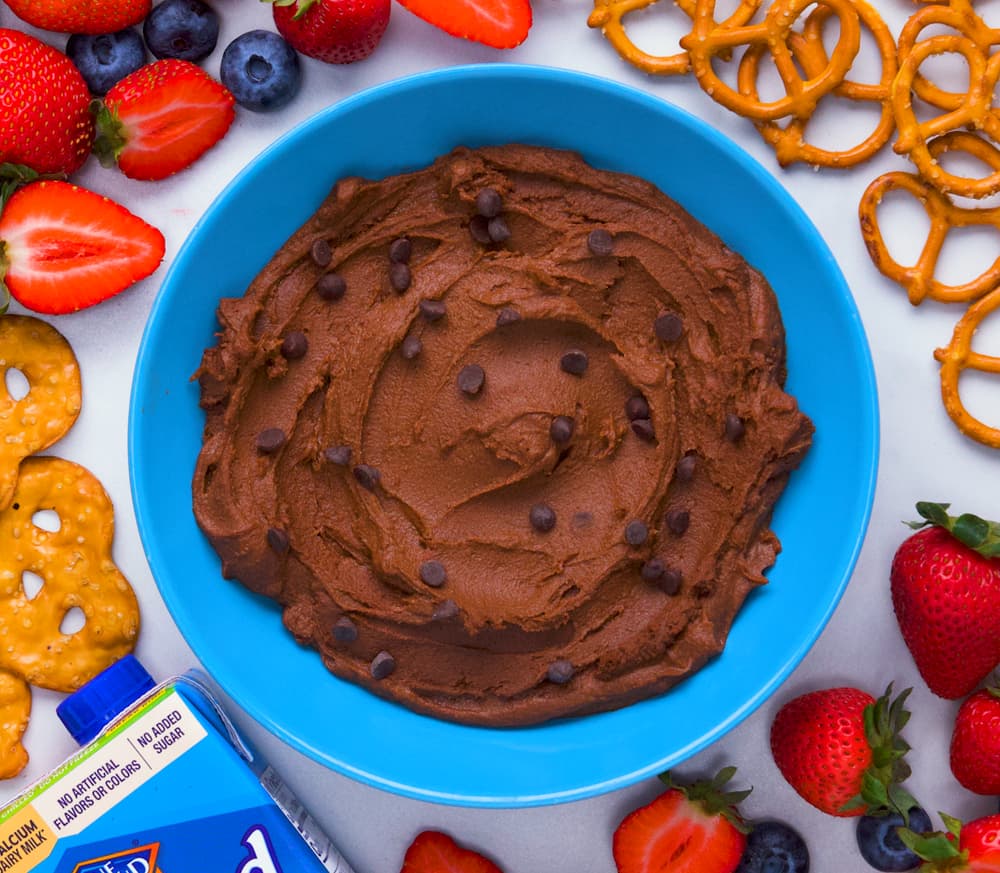 A blue bowl of chocolate dip with chocolate chips, surrounded by strawberries, blueberries, pretzels, and a carton of almond milk.