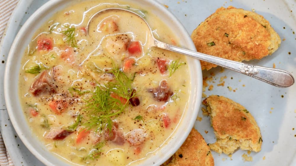 Bowl of creamy soup with vegetables and herbs, served with two pieces of cornbread on a blue plate, spoon resting in the soup.