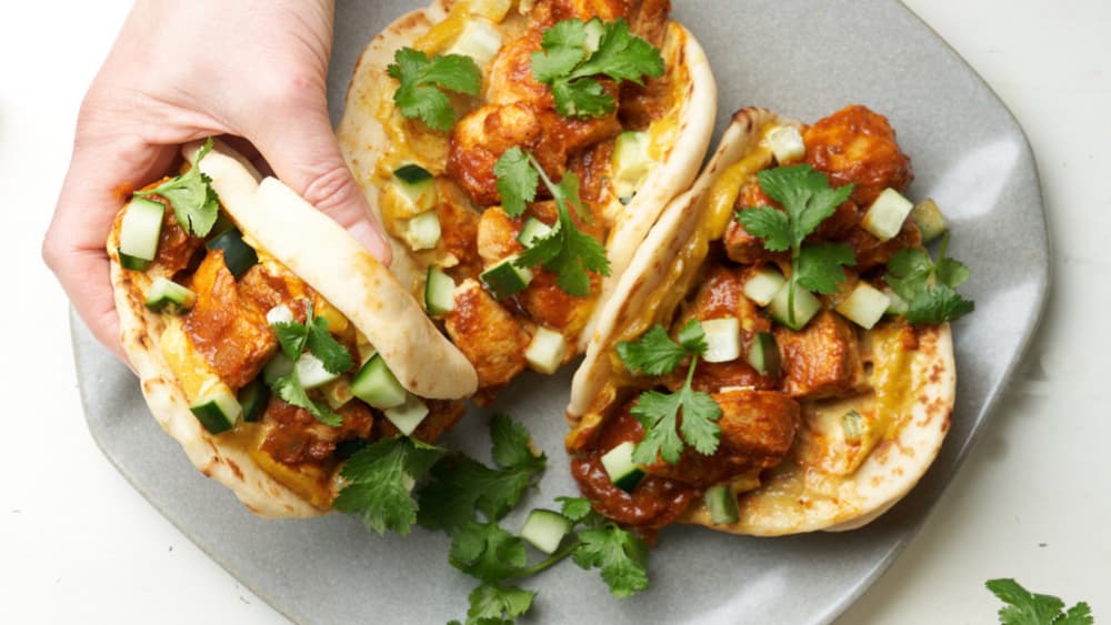 Three flatbreads topped with spiced chicken, diced cucumber, and fresh cilantro on a gray plate; a hand is picking up one flatbread.