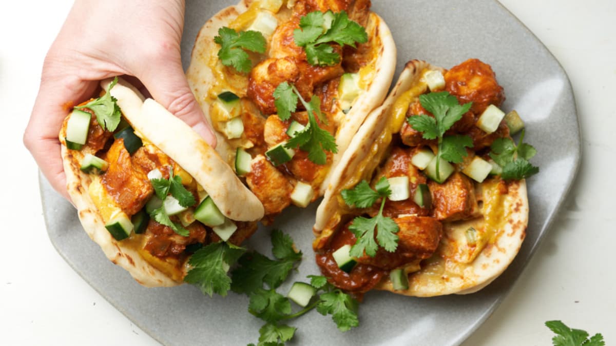 Three flatbreads topped with spiced chicken, diced cucumber, and fresh cilantro on a gray plate; a hand is picking up one flatbread.