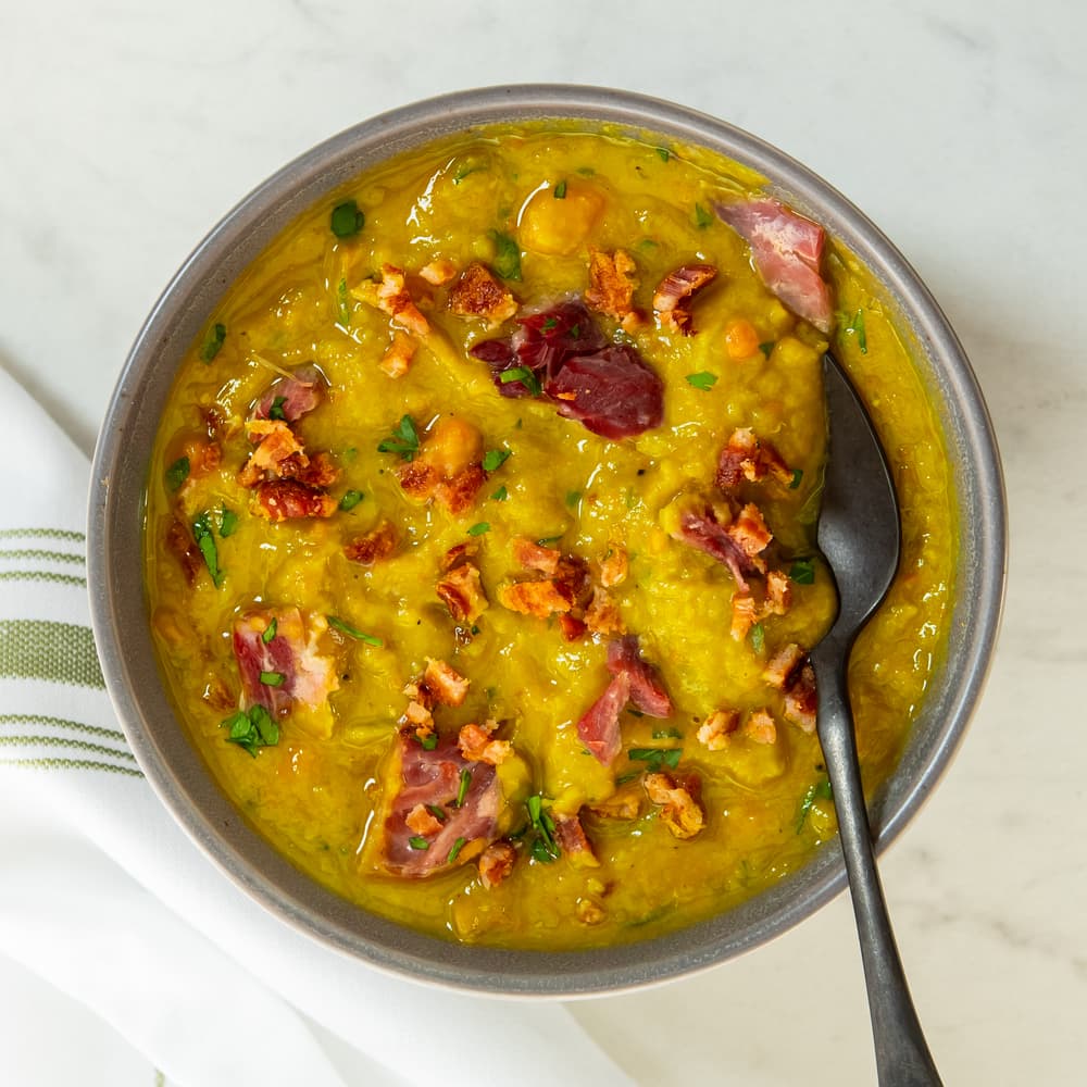 Bowl of thick yellow split pea soup with pieces of ham and herbs, placed on a white surface with a spoon and a striped cloth napkin beside it.
