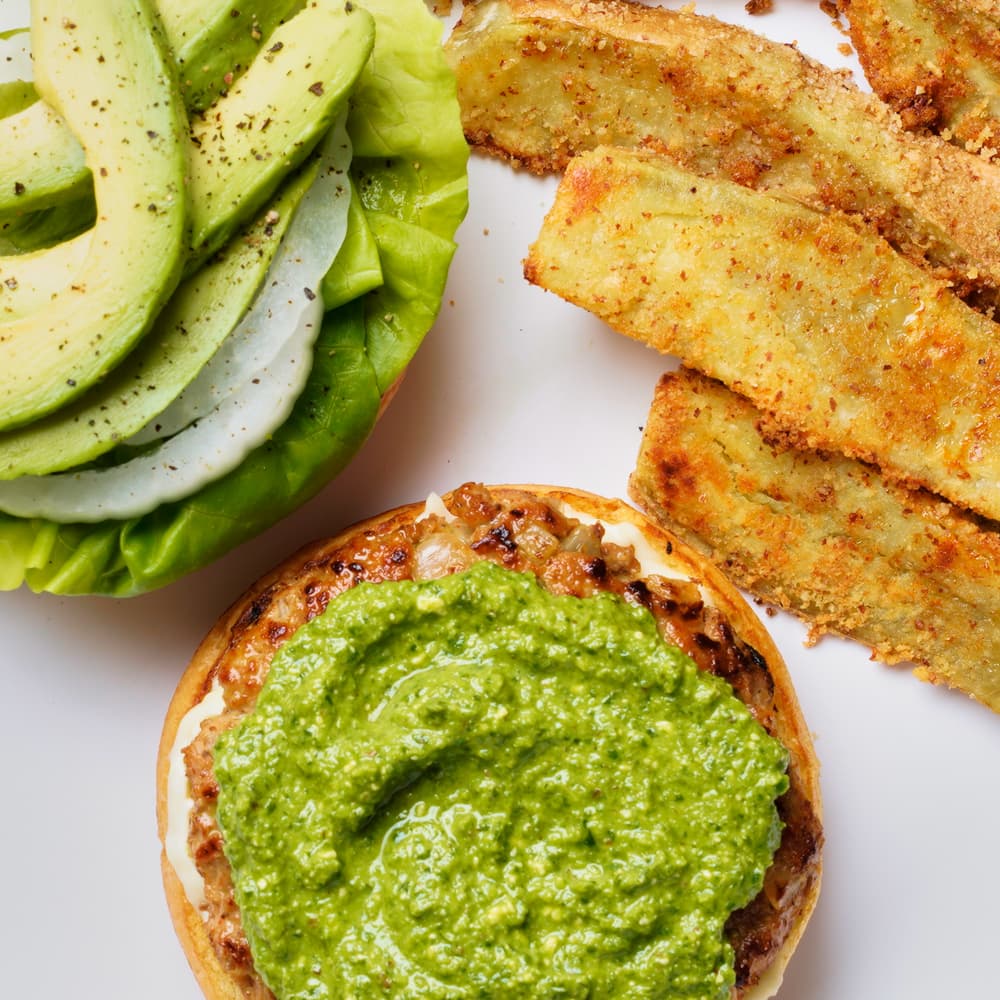 Open burger bun with green sauce, leaf lettuce, onion, and avocado slices beside breaded, baked potato wedges on a white plate.