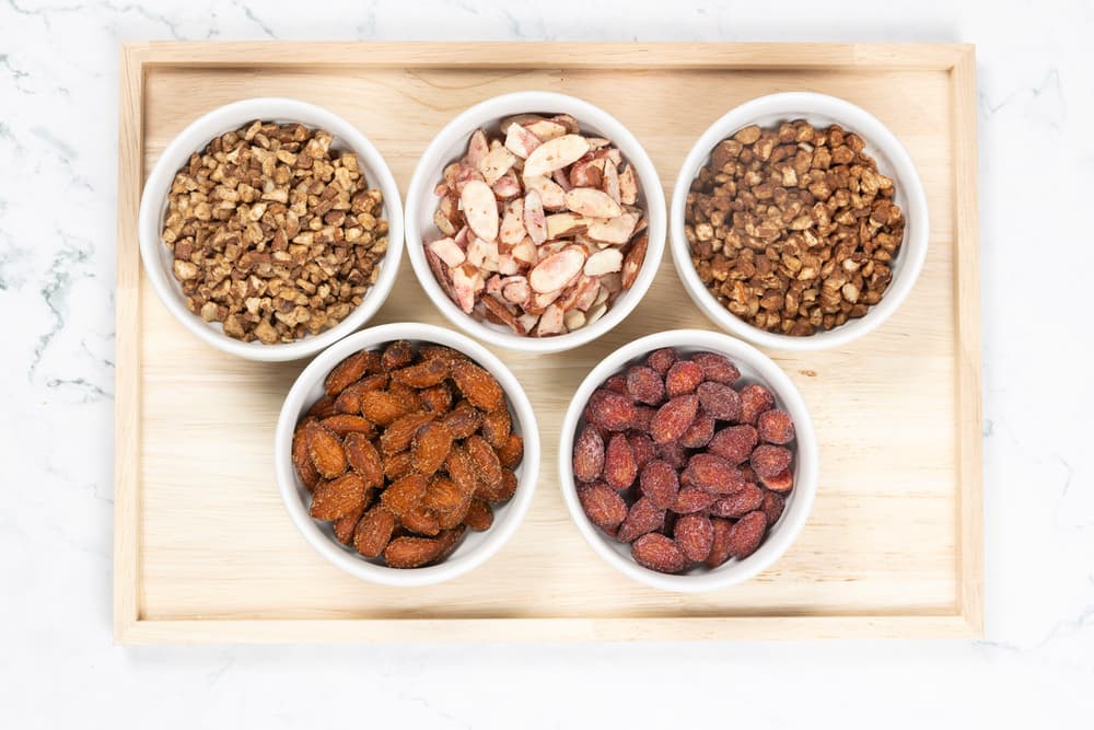 Five small bowls with assorted nuts, including almonds, Brazil nuts, and chopped varieties, are arranged on a wooden tray on a white surface.