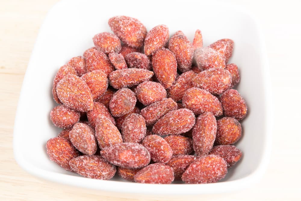 A white bowl filled with red, sugar-coated almonds sits on a light wooden surface.