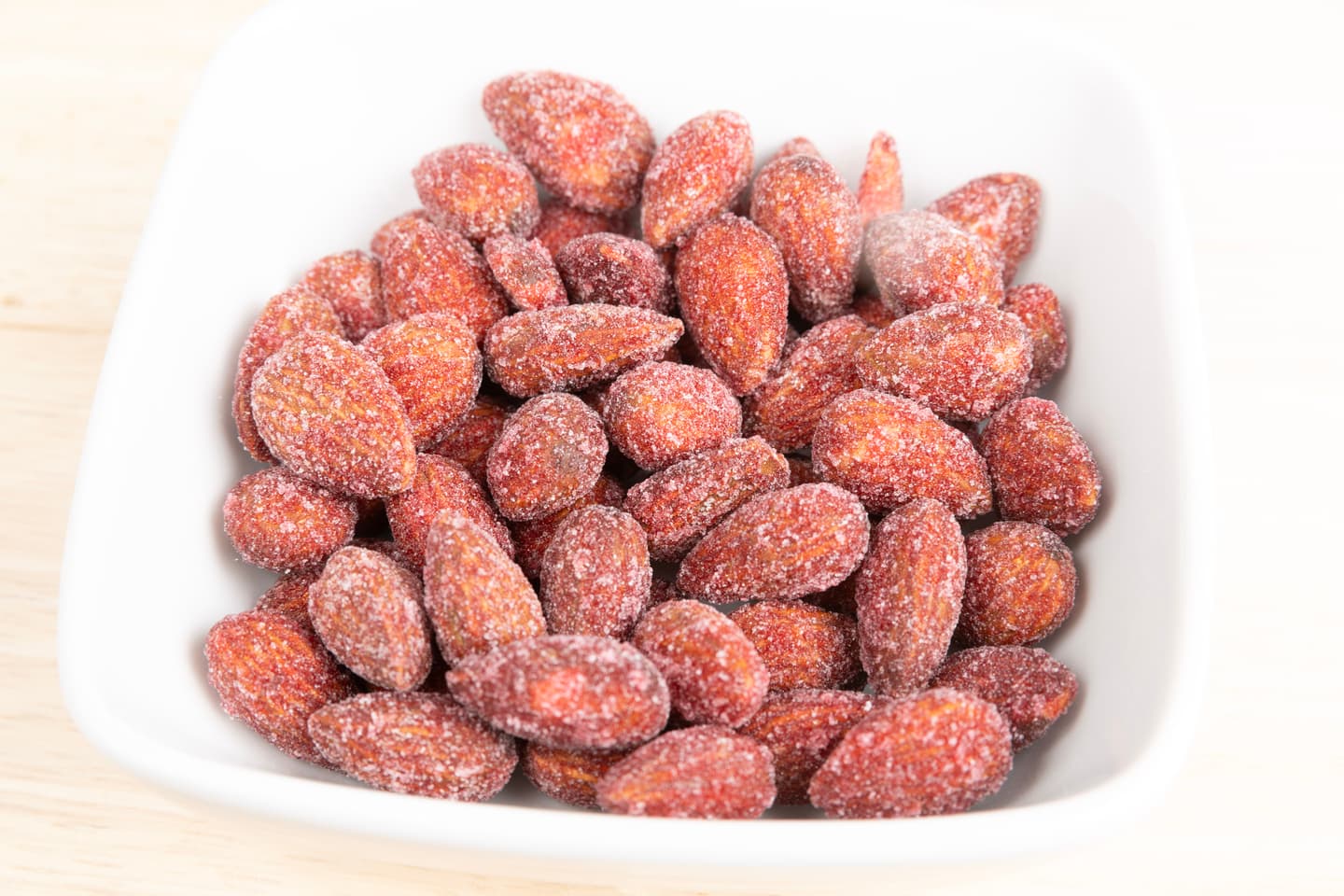 A white bowl filled with red, sugar-coated almonds sits on a light wooden surface.