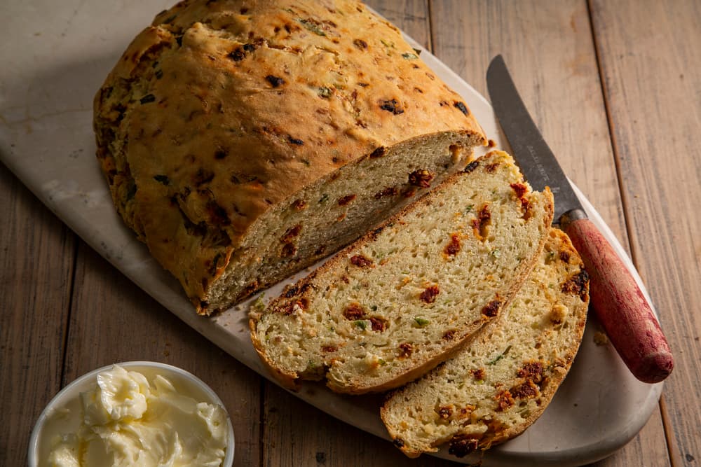 Sliced savory bread with herbs and sun-dried tomatoes on a cutting board, a knife, and a small bowl of butter on a wooden table.