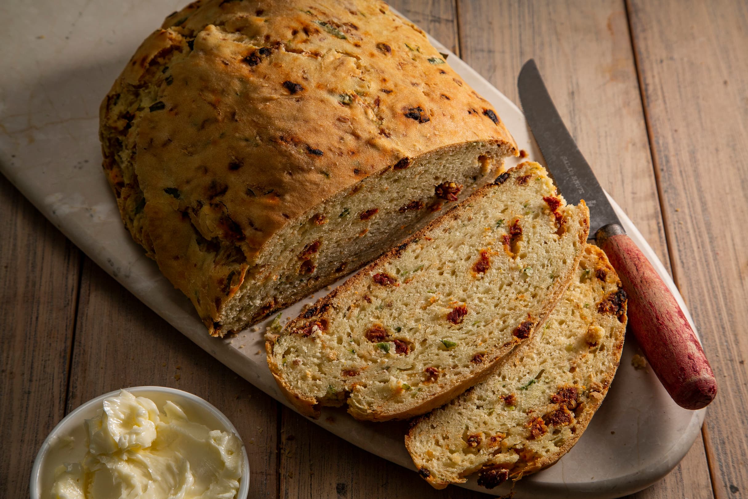 Sliced savory bread with herbs and sun-dried tomatoes on a cutting board, a knife, and a small bowl of butter on a wooden table.