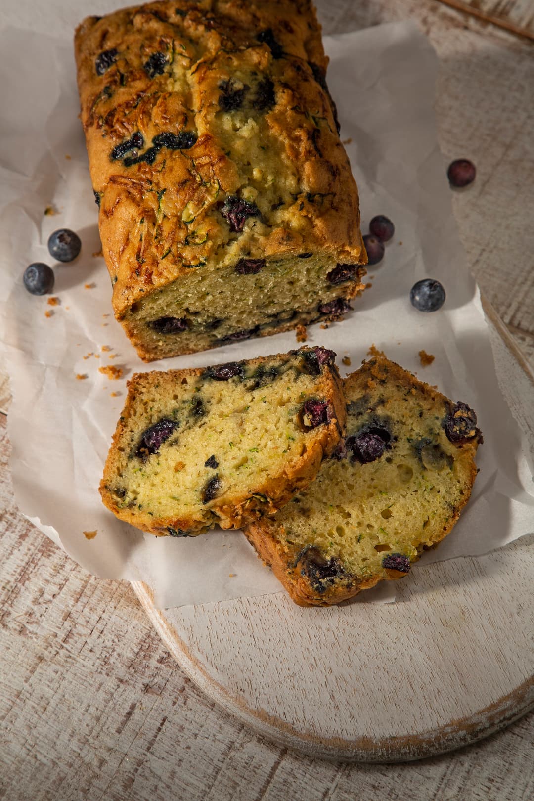 A loaf of blueberry bread with two slices cut, resting on parchment paper atop a wooden board, with a few loose blueberries nearby.
